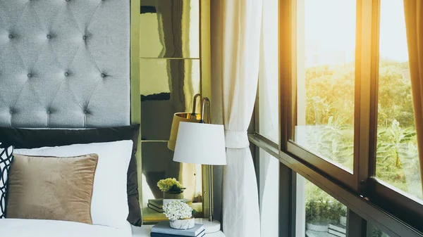 modern bedroom beside window in the morning with sunlight, pillows and cushions in white, beige and brown color tone on bed in luxury bedroom at home