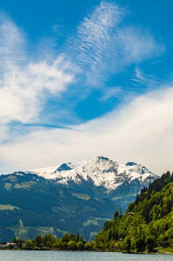 Göl, yeşil ağaçlar ve mavi bulutlu gökyüzünün altındaki karlı kayalar.