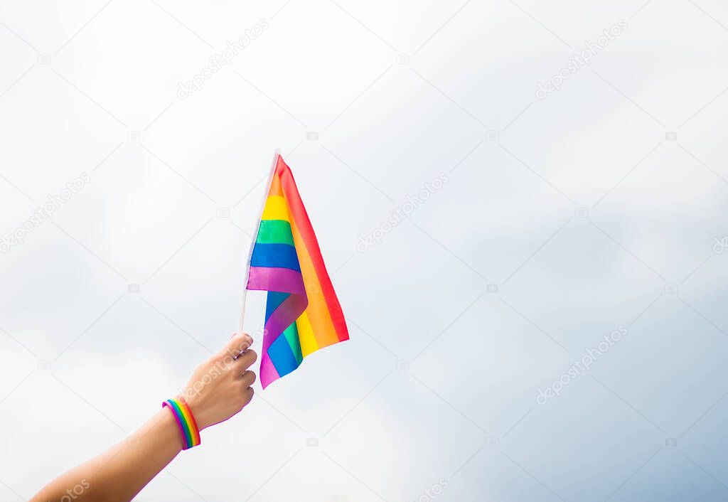  hand wearing a gay pride bracelet with rainbow flags against the sky. LGBT people, same-sex relationships and homosexual concepts 