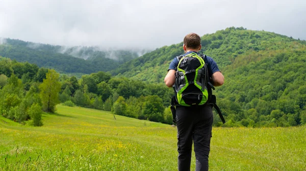 Man with green backpack hiking. Meadow overlooking the mountains and fog. Low wooded mountains. Summer weekend.