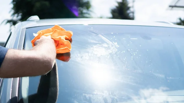 Mão limpando o pára-brisas de um carro em um dia ensolarado. Limpe com um pano laranja. Rag limpa manchas de água na janela — Fotografia de Stock