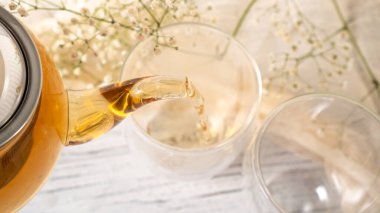 Şeffaf cam çaydanlıktan cam fincana altın çay dökülüyor. Bardağa siyah çay dolduran cam çaydanlık. Beyaz ahşap masa