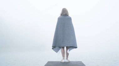 Ilık gri bir battaniyeye sarılı yalnız genç kız gölün yakınındaki tahta bir uçurumda duruyor ve sise bakıyor.