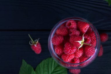 Koyu arkaplanda yeşil yapraklı kırmızı taze ahududu. Tahta bir masada meyveli doğal olgun organik böğürtlenli cam bardak..