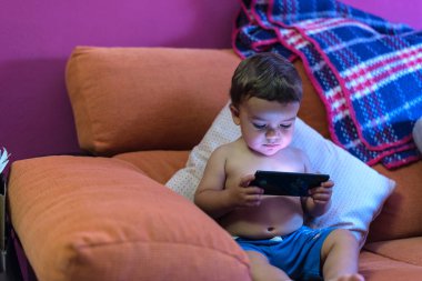 Küçük çocuk koltukta oturmuş cep telefonuyla video izliyor.