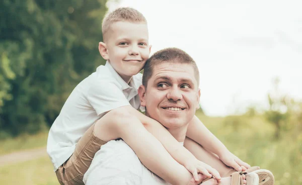 Dad and son are walking in nature, dad is holding son on his shoulders and son is smiling