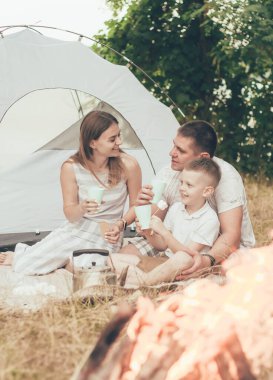 Çadırın yakınındaki bir kamp ateşinde oturan, kurabiye yiyen ve çay içen aile.