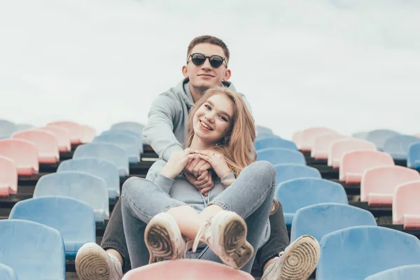 A guy hugs a girl, a guy and a girl in identical sweatshirts are sitting in a stadium