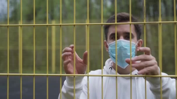 Uomo mascherato rinchiuso dietro le sbarre in quarantena durante la pandemia. Un uomo si avvicina alla recinzione e si guarda intorno — Video Stock