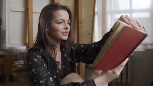 Mladý krásný umělec dívka čte knihu v ateliéru umělců v blízkosti stojanu a usmívá — Stock video