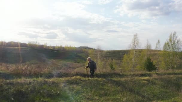 Старый седой мужчина турист с рюкзаком и тростью в руке, спускающийся по холмам во время заката, кадры с воздуха — стоковое видео