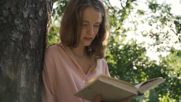 Dívka v růžové košili čte knihu v lese poblíž stromu za slunečného dne — Stock video