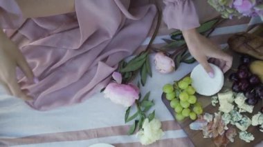 Pembe elbiseli bir kız yazın bir piknikte yemek hazırlar. Peynir, üzüm, kiraz, fındık, çiçek.