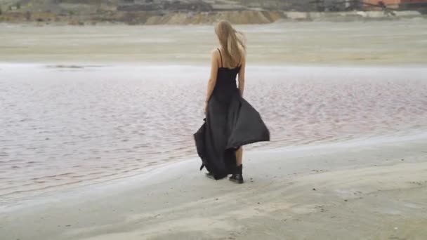 Uma menina em um vestido preto caminha para a água em terras contaminadas em meio a uma planta de processamento de cobre — Vídeo de Stock