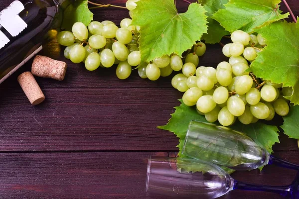 A bunch of green grapes, a bottle of wine and wine glasses on wooden background. Top view, flat lay, copy space.