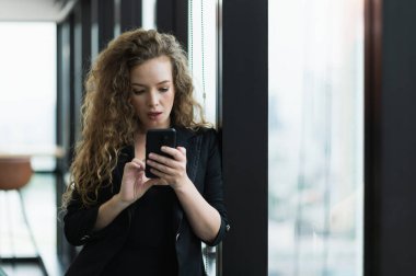 Ofiste pencerenin yanında duran akıllı telefonu kullanan güzel seksi kadın..