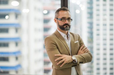 İş yerinde kolları kavuşturulmuş, pencerenin yanında duran yakışıklı iş sakallı genç adam..