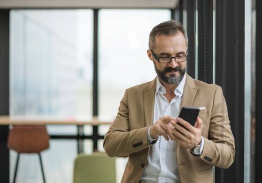 Genç yakışıklı iş sakallı adam ofis yerinde akıllı telefon kullanarak pencerenin yanında dikiliyor..