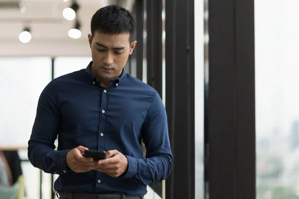 Business asian man using smartphone standing near window in office.