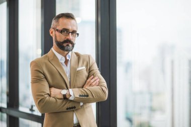 İş yerinde kolları kavuşturulmuş, pencerenin yanında duran yakışıklı iş sakallı genç adam..