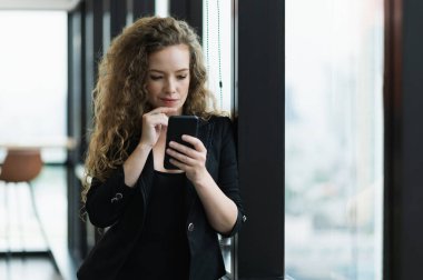 Ofiste pencerenin yanında duran akıllı telefonu kullanan güzel seksi kadın..