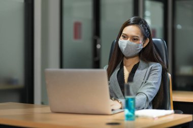 Genç Asyalı iş kadını, modern ofiste dizüstü bilgisayar kullanıyor. Masaya maske jeli sürerek sosyal mesafelerle çalışıyor..