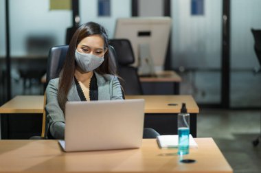 Genç Asyalı iş kadını, modern ofiste dizüstü bilgisayar kullanıyor. Masaya maske jeli sürerek sosyal mesafelerle çalışıyor..