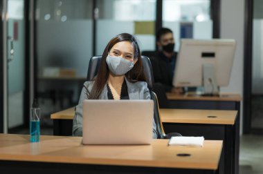 Genç Asyalı iş kadını, modern ofiste dizüstü bilgisayar kullanıyor. Masaya maske jeli sürerek sosyal mesafelerle çalışıyor..