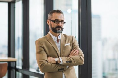 İş yerinde kolları kavuşturulmuş, pencerenin yanında duran yakışıklı iş sakallı genç adam..