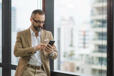 Genç yakışıklı iş sakallı adam ofis yerinde akıllı telefon kullanarak pencerenin yanında dikiliyor..