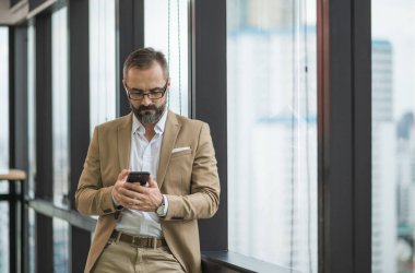 Genç yakışıklı iş sakallı adam ofis yerinde akıllı telefon kullanarak pencerenin yanında dikiliyor..