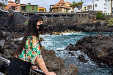 Korkuluğa yaslanmış genç bir Asyalı kadın. Çiçekli bir gömlek ve maske takıyor. San Telmo bulvarında, Puerto de La Cruz 'da..