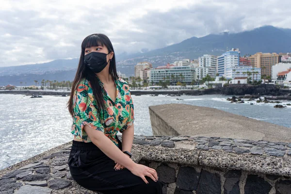 Genç Asyalı turist oturmuş denizi seyrediyor. Çiçekli bir gömlek ve maske giy. Puerto De la Cruz, Tenerife 'yi gezmek.