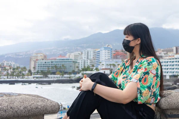 Genç Asyalı turist oturmuş denizi seyrediyor. Çiçekli bir gömlek ve maske giy. Puerto De la Cruz, Tenerife 'yi gezmek.