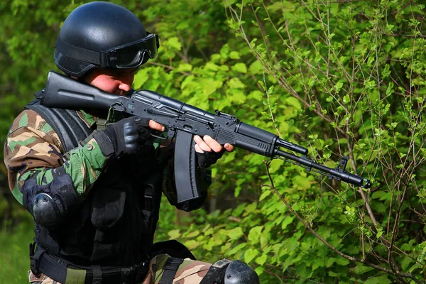 Airsoft soldier in a helmet and with a gun on a blurred background of trees.