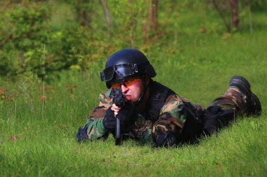 Kasklı ve silahlı bir adam yeşil çimlerin üzerinde, ormanda bir yerden ateş ediyor..