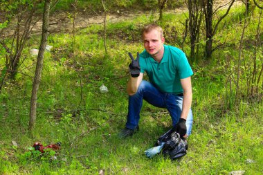 Marrs yeşili tişörtlü gönüllü bir genç çöp torbası doğada çözünemeyen plastik çöpleri topluyor..