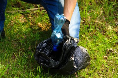 Marrs yeşili tişörtlü gönüllü bir genç çöp torbası doğada çözünemeyen plastik çöpleri topluyor..