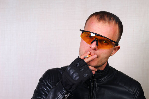 A happy handsome young biker in a black leather jacket and goggles is smoking a cigarette.