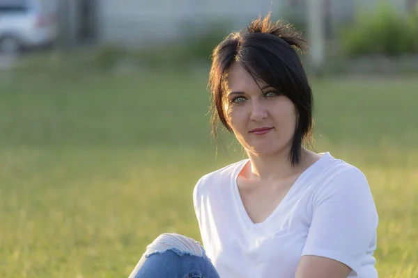 若いです女性で白いTシャツとフリップジーンズに芝生の上に座ってぼやけた背景 — ストック写真