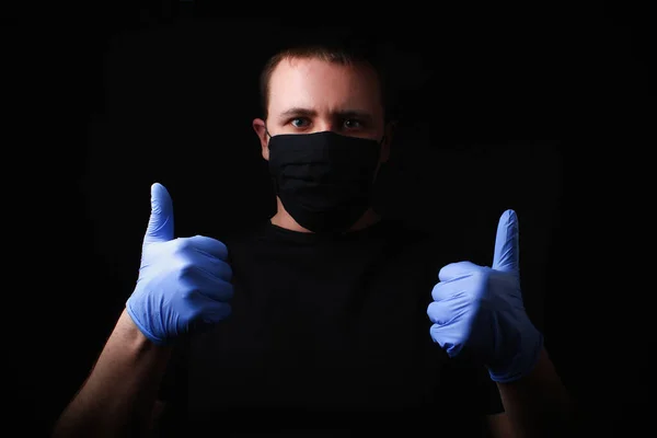 A man in a black mask and medical gloves shot on a black background in a dark or low key. Holds thumbs up, concept - everything will be fine