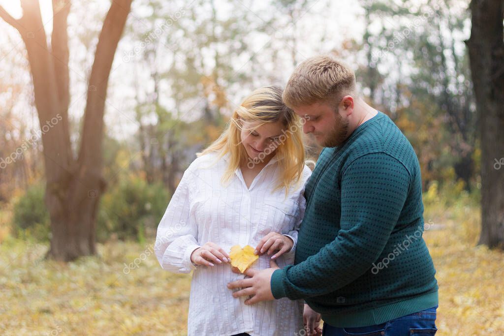 A pregnant woman holds a yellow leaf in the shape of a heart as a symbol of a new heart, and a caring husband touches her tummy, toned