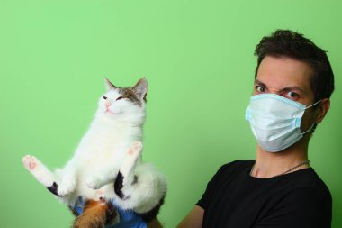 Cerrahi maske takmış bir adamın elindeki kedi. Yeşil arka plan Sevgi ve evcil hayvan bakımı kavramı