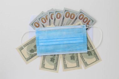 Beyaz ahşap bir arka planda klasik bir tıbbi cerrahi maskeyle kaplanmış Amerikan doları. Kriz Covid-19 temasından kaynaklanıyor