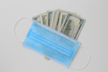 Beyaz ahşap bir arka planda klasik bir tıbbi cerrahi maskeyle kaplanmış Amerikan doları. Kriz Covid-19 temasından kaynaklanıyor