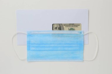 İçinde dolar olan klasik beyaz bir posta zarfı ve beyaz ahşap bir arka planda tıbbi maske.