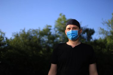 Günlük giysiler ve tıbbi maske takmış beyaz bir genç ya da yetişkin bir adam şehir parkında dinleniyor..