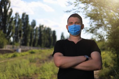 Günlük giysiler ve tıbbi maske takmış beyaz bir genç ya da yetişkin bir adam şehir parkında dinleniyor..