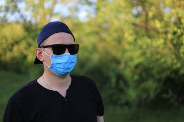 Günlük giysiler ve tıbbi maske takmış beyaz bir genç ya da yetişkin bir adam şehir parkında dinleniyor..