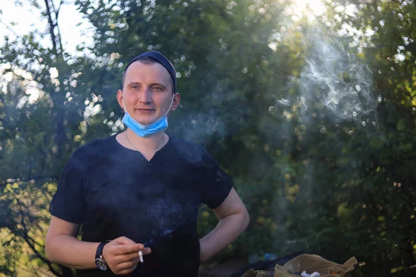 A young white man or a young adult Caucasian guy smokes a cigarette with a medical surgical mask on his chin.